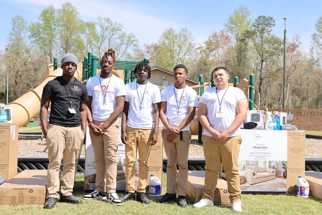 Five people dressed in t-shirts and brown pants standing in a park with boxes of furniture around them.