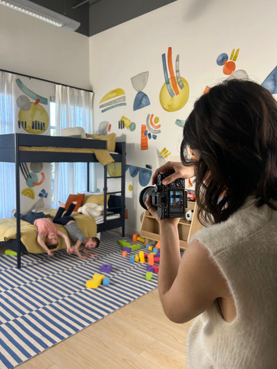 Person taking a photo of a child playing on a bed in a room with colorful wall art.