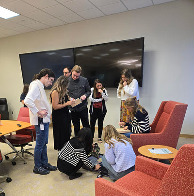 Group of people gathered around a table in a modern office setting with red chairs and large monitors on the wall.